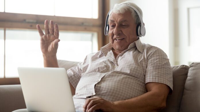 Smiling senior man wear earphones wave to camera having video call on laptop